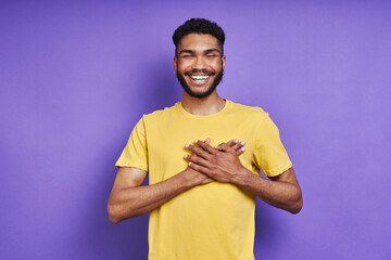Thankful African man holding hands on heart and smiling while standing against purple background