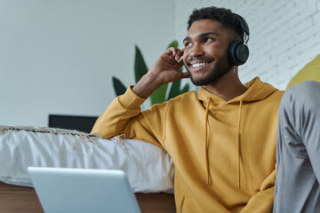 Handsome African man in headphones using laptop while sitting on the floor near bed at home