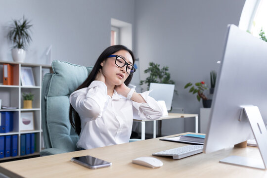 Young beautiful Asian woman office worker, freelancer, business woman in glasses and white shirt, sitting at the table at the computer, tired, exhausted, doing neck massage with two hands
