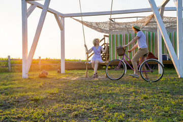 young couple in love, woman is sitting on the swing, man is riding bicycle on the lavender field, sunset