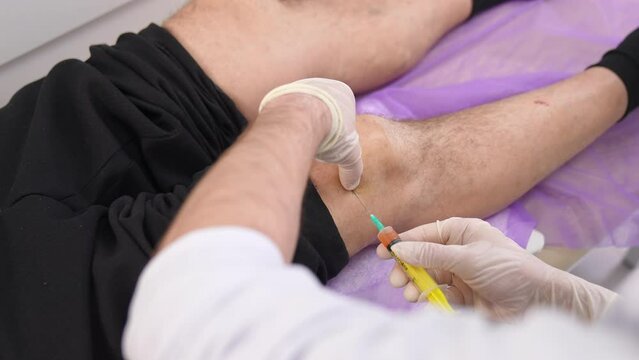 Injection of medicines into a knee. Gloved hands make a shot to a patient&rsquo;s leg lying on the bed. Top view.