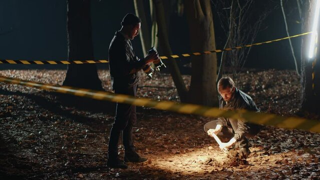 Two Caucasian detectives with shovel in hands digging hole, talking to each others in forest at night. Footage of young smart men at work. Mysterious murder. Strange story. Horror scene