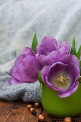 Vertical photo of purple tulip buds in a green mug on a wooden table close-up with a background of light fabric