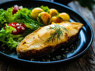 Fish dish - fried halibut with fried potatoes, cherry tomatoes and leafy greens on wooden table
