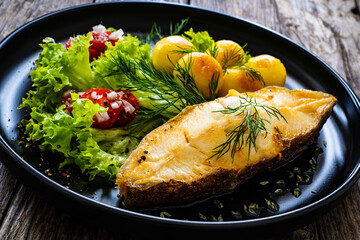 Fish dish - fried halibut with fried potatoes, cherry tomatoes and leafy greens on wooden table
 - obrazy, fototapety, plakaty