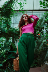 Obraz na płótnie Canvas Fashionable woman wearing pink asymmetric ruffled blouse, green trousers, holding straw wicker handbag, walking in tropical garden. Summer fashion conception