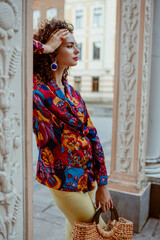 Summer fashion portrait of elegant curly brunette woman wearing trendy colorful printed shirt, holding wicker straw top handle bag, posing in street of the city
