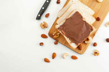 Square bread for toast with chocolate paste on a wooden board. Nuts, a knife and a wooden cutting board with a sandwich and slices of bread on a white table.