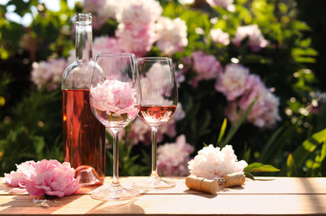 Bottle and glasses of rose wine near beautiful peonies on wooden table in garden