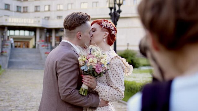 professional wedding photographer takes photographs of beautiful bride and groom couple kissing in park. man in gray suit hugs woman with red hair in white dress.