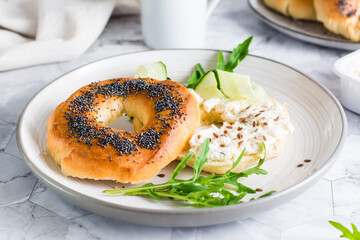 Bagel sandwich with poppy seeds with ricotta, cucumber and arugula on a plate. Homemade breakfast