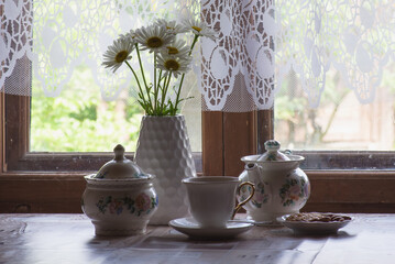 a tea set and a vase with daisies on the table in front of a wooden window of a rural house