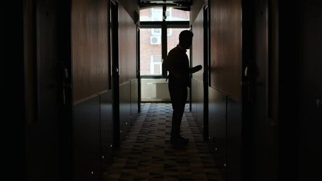 silhouette of unrecognizable man enter hotel room in hallway. businessman in trousers and vest walks to door along dark corridor, behind window creates light. concept of business trip