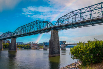 Fototapeta na wymiar the Walnut Street Bridge over the rippling blue waters of the Tennessee River surrounded by lush green trees and buildings with blue sky, clouds and a rainbow at Coolidge park in Chattanooga Tennessee