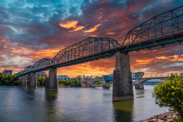 the majestic Walnut Street Bridge over the rippling blue waters of the Tennessee River surrounded by lush green trees and buildings with powerful clouds at sunset at Coolidge park in Chattanooga - obrazy, fototapety, plakaty