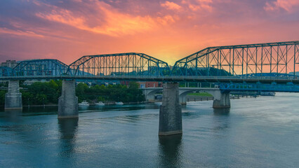 Fototapeta na wymiar an aerial shot of the Walnut Street Bridge over the rippling blue waters of the Tennessee River surrounded by lush green trees and buildings with powerful clouds at sunset at Coolidge park