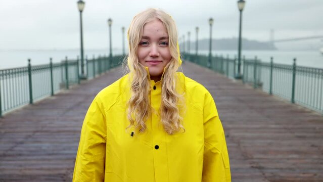 Woman wearing the hood during a rainy day by the ocean. Portrait of a blonde, joyful girl in a yellow raincoat with the Golden Gate Bridge in the background. High quality 4k footage