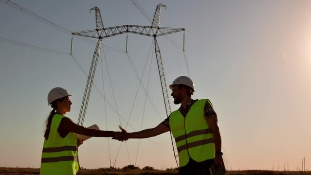 Two electrical engineers meet and shake hands against the backdrop of high-voltage electrical tower at sunset. Meeting of workers near power grids. Teamwork, signing contract, two businessmen
