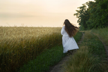 A girl with long blond hair walks through a field of ripe wheat in a white long robe