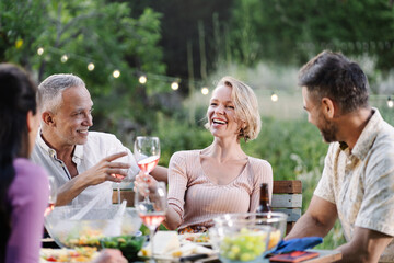 Mature friends having dinner around table in a garden on a summer evening, drinking wine, having...