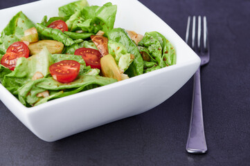 white bowl with fresh organic lettuce, and tomatoes and cucumbers, next to it is a fork, on gray kitchen counter