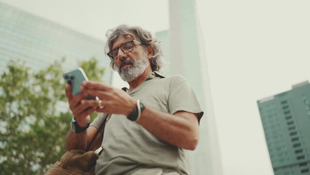 Friendly middle aged man with gray hair and beard looking at map trying to find his way using his mobile phone. Mature gentleman in eyeglasses using map app in cellphone outdoors