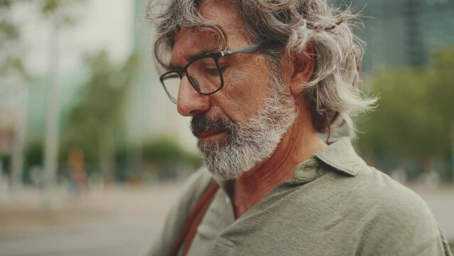 Clouse-up, friendly middle aged man with gray hair and beard looking at map trying to find his way using his mobile phone. Mature gentleman in eyeglasses using map app in cellphone outdoors