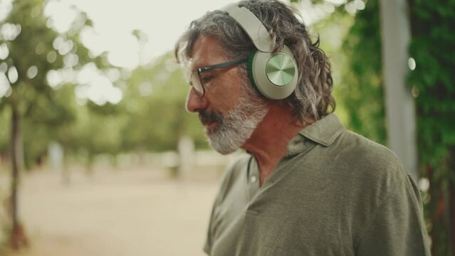 Clouse-up, friendly middle-aged man with gray hair and beard wearing casual clothes listening to music on headphones. Mature gentleman in eyeglasses enjoys music outdoors
