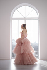 A girl in a beautiful lush long dress of dusty rose color stands near an open window. Vintage...