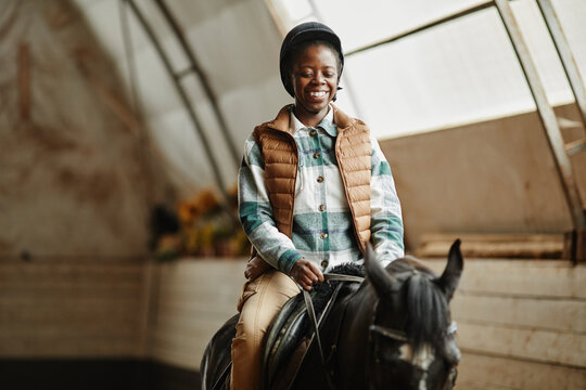 Portrait of smiling African American woman riding horse in indoor arena at horse ranch or practice stadium, copy space