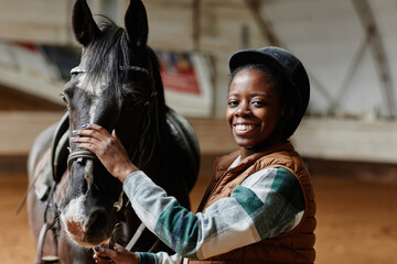 Waist up portrait of smiling black woman posing with horse in indoor riding arena and looking at...