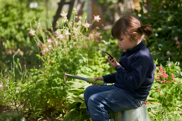 A girl in the garden near the flower garden with a smartphone in one hand and a small rake in the other hand. Selective focus.