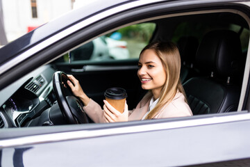 Obraz na płótnie Canvas Beautiful woman is drinking coffee, looking at camera and smiling while sitting in her car