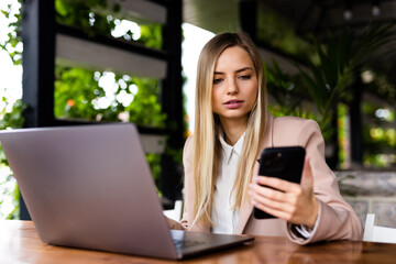 Young woman sitting in coffee shop at wooden table, drinking coffee and using phone on table is laptop.