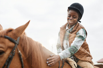 Warm toned portrait of young black woman riding horse in sunlight and smiling while bonding with...