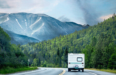 Retro camper driving on highway sucrrounded by evergreen forests and tall mountains with snow...