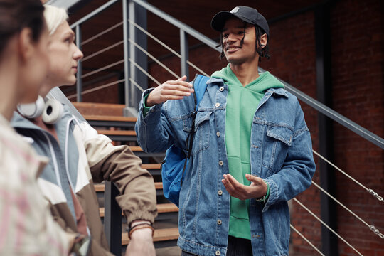 Young smiling black man in casualwear standing on staircase by brick wall of building and chatting with his colleage friends after lessons