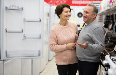 Obraz premium European spouses of mature age, who have come to the electronics and household appliances store for shopping, choose a ..refrigerator, examining it