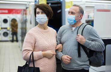 Obraz premium Senior couple in face masks standing in salesroom of tech store while shopping.