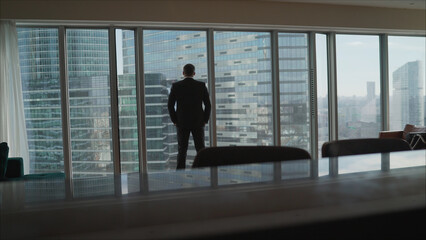 Fototapeta na wymiar A man in a black suit stands at a panoramic window overlooking skyscrapers. One Young Man Standing on Office Balcony Overlooking City Skyline.