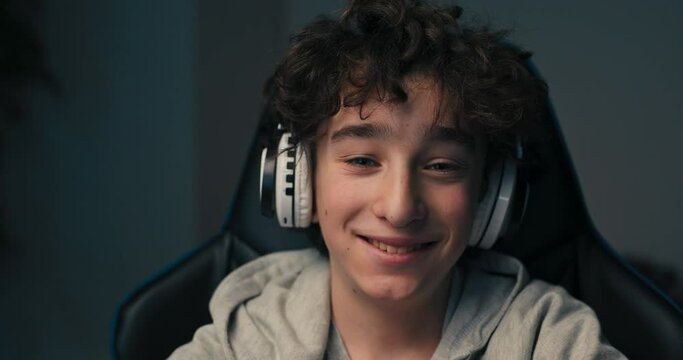 Face portrait of a boy with curly long hair wearing a sweatshirt. The child is sitting in a chair in front of a computer, wearing wireless headphones.