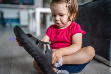 one caucasian toddler girl child playing with slippers at home in room alone putting on her feet...