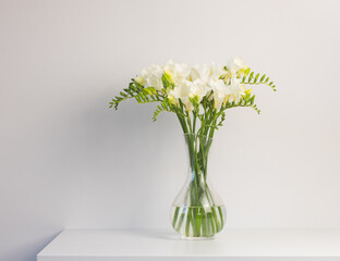 Close up of white freesia flowers in glass vase on edge of table against plain background (selective focus)