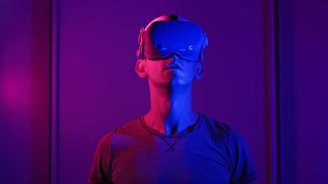 Young man watches 360 media content in VR Helmet. Male interact with virtual reality using VR headset at red-blue light. Futuristic technology. Immersion in meta-universe, augmented reality, metaverse