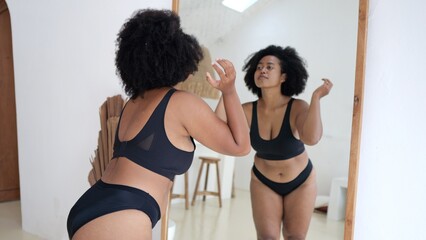 Multicultural plus size woman in lingerie looking at her body in the mirror. Black woman struggles with obesity and overweight, wants to lose weight and be slim Woman body positive without criticism - obrazy, fototapety, plakaty