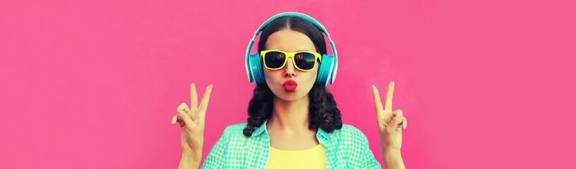 Portrait of stylish woman listening to music with headphones blowing her lips sending sweet air kiss wearing sunglasses on pink background