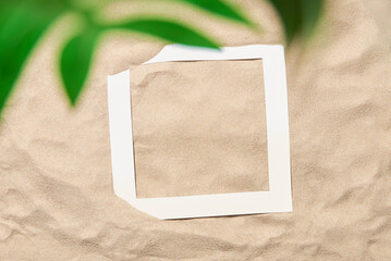 Tropical composition, concepts of summer, beach and vacations: smooth sand background with an empty paper frame and green leaves peeking out in the foreground. Minimal creative design with copyspace.