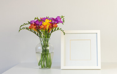 Close up of colourful freesia flowers in glass vase with blank square picture frame against white wall (selective focus)