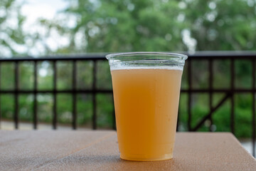 A tall clear plastic glass of sour craft beer in a microbrewery. The cold refreshment has frost on the glass. The beer is on a wooden table. The top of the glass has white froth around it. 