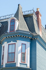 The top exterior corner roof of a historical Victorian building is made of blue clapboard, white and red trim vintage windows, and a large turret in the corner of the decorative heritage building. 